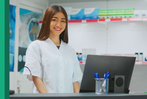 Jovem farmacêutica sorridente com cabelos longos e pretos em ambiente de farmácia, à sua frente uma tela de computador