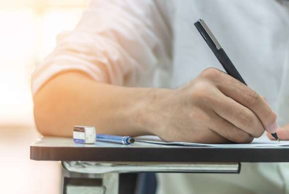 Foto de homem jovem branco destro sentado em uma cadeira estudantil, vestindo camisa branca segurando uma caneta e escrevendo sobre uma folha. A mesa de madeira também contém uma borracha e uma lapiseira. O fundo é desfocado.