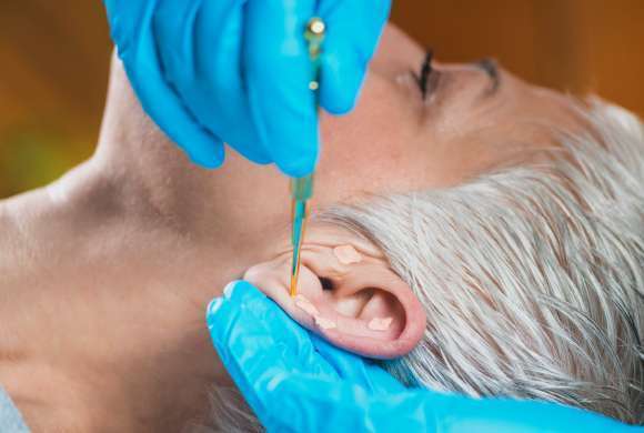 Mulher de cabelo curto branco deitada recebe tratamento na orelha feito por pessoa com luvas azuis segurando umapinça 