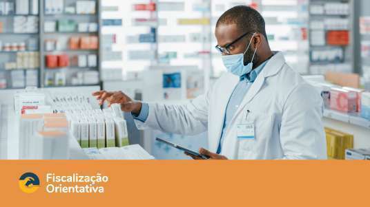 Farmacêutico negro, de óculos, jaleco branco e máscara confere medicamentos em uma prateleira dentro da farmácia. Abaixo uma tarja laranja escrita Fiscalização orientativa 
