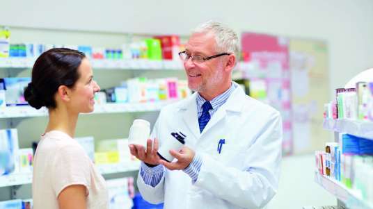 Farmacêutico grisalho e de óculos de jaleco branco conversa com mulher de camise ta clara e cabelo preso, ambos estão em pé dentro da farmácia  